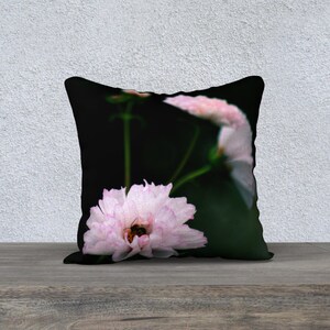 May include: A decorative pillow featuring a close-up of a light pink flower with a bee inside. The flower has multiple petals with a darker pink edge. The background is dark green, and the pillow is on a wooden surface.