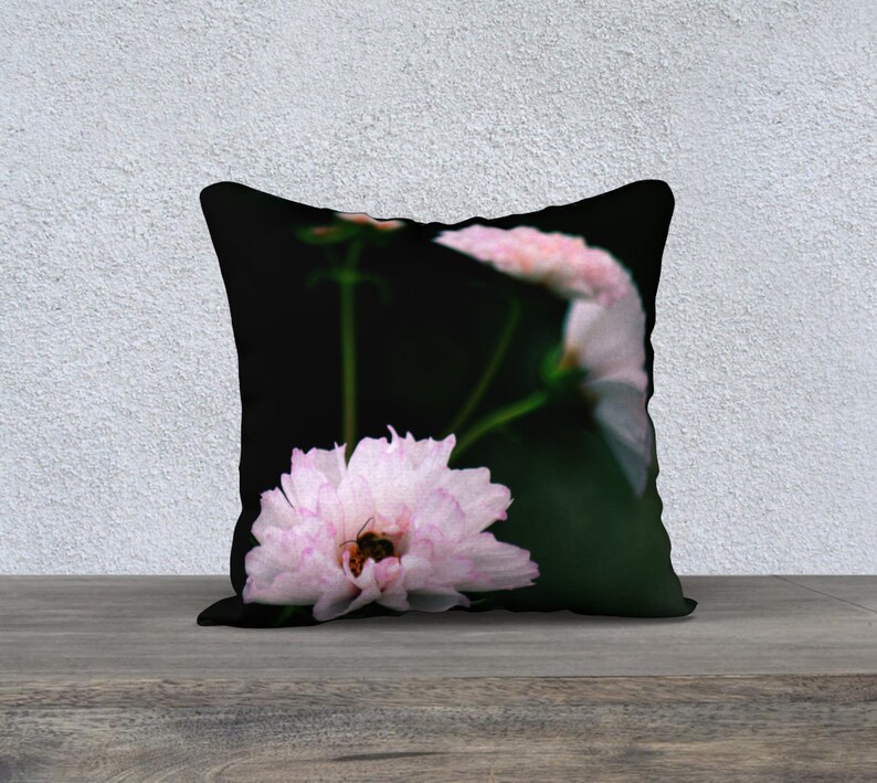 May include: A decorative pillow featuring a close-up of a light pink flower with a bee inside. The flower has multiple petals with a darker pink edge. The background is dark green, and the pillow is on a wooden surface.