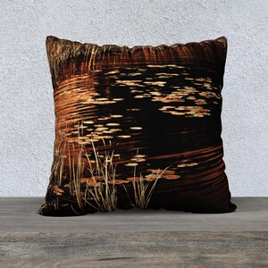 May include: A decorative throw pillow with a dark brown background featuring a pattern of golden lily pads and reeds in a pond.