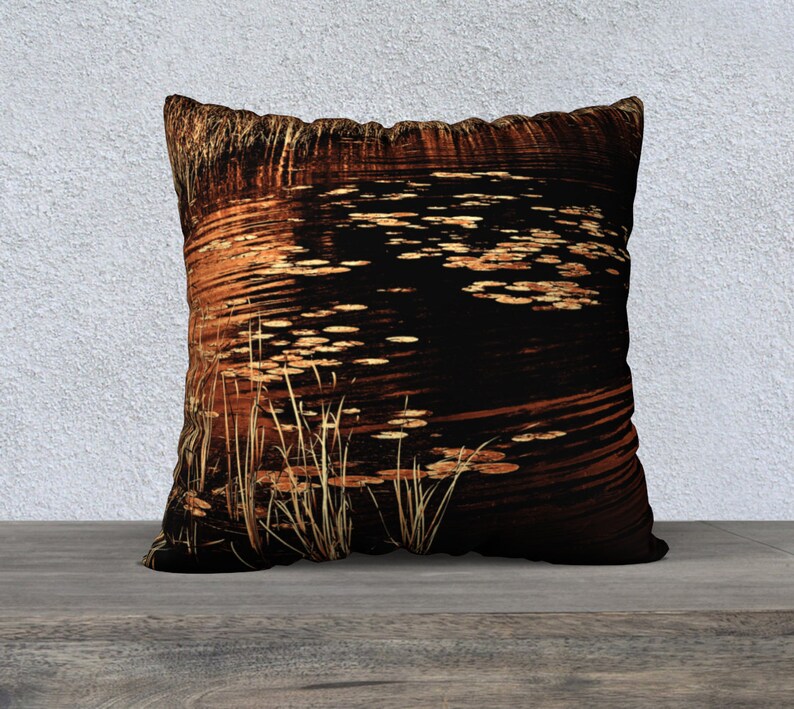 May include: A decorative throw pillow with a dark brown background featuring a pattern of golden lily pads and reeds in a pond.