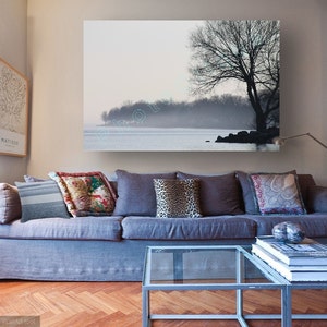 May include: A gray sofa with pillows in a living room setting. A large framed print of a tree and a lake hangs on the wall above the sofa. A glass coffee table sits in front of the sofa with a stack of books on top.