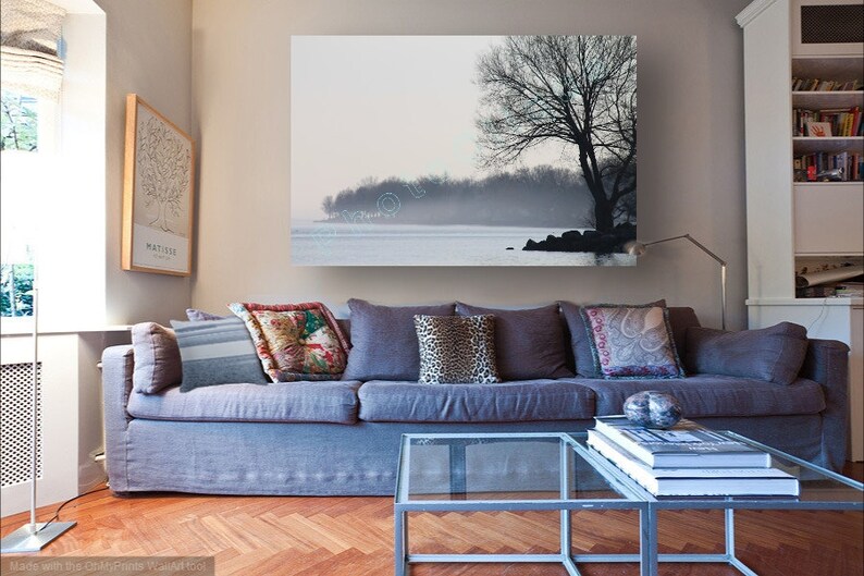 May include: A gray sofa with pillows in a living room setting. A large framed print of a tree and a lake hangs on the wall above the sofa. A glass coffee table sits in front of the sofa with a stack of books on top.
