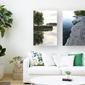 May include: Two framed landscape photographs of a lake with a tree and a rocky shoreline. The photographs are hung on a white wall above a white couch.