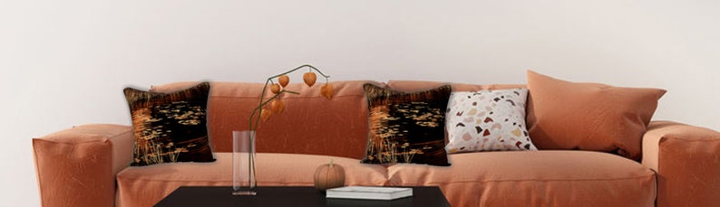 May include: A burnt orange sofa with three throw pillows. The pillows have black and gold geometric patterns, a white and brown geometric pattern, and a solid burnt orange color. A vase with dried branches and a small pumpkin are on the coffee table in front of the sofa.