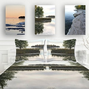 May include: A bedroom with a white bedspread and two pillows featuring a lake scene. Three framed pictures are displayed on the wall above the bed, each showcasing a different lake scene. A vase with branches is positioned on the right side of the bed.