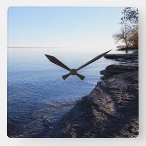 Puede incluir: Un reloj cuadrado con una esfera y agujas negras. La imagen de fondo es un lago azul con una costa rocosa y un cielo azul.