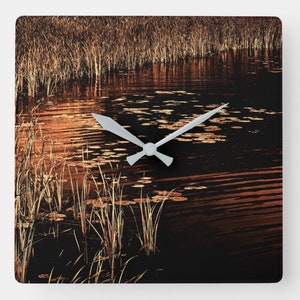 Puede incluir: Un reloj cuadrado con una esfera blanca y agujas negras. La esfera del reloj está superpuesta con una fotografía de un estanque tranquilo con juncos y nenúfares. El agua es de color marrón oscuro con reflejos naranja y amarillos.