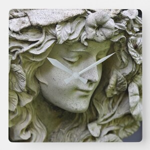 May include: A white stone sculpture of a woman's face with a clock face superimposed over her eyes. The clock hands are white and the sculpture is surrounded by leaves.