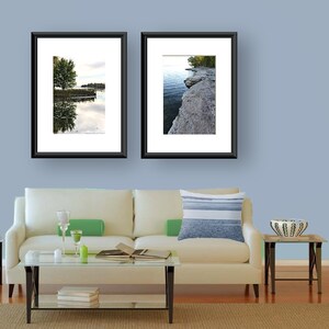 May include: Two framed landscape photographs of a lake and shoreline with trees hanging over the water. The photos are hanging on a light blue wall above a white couch with a blue and white striped pillow.