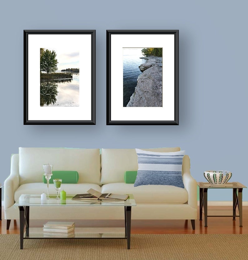 May include: Two framed landscape photographs of a lake and shoreline with trees hanging over the water. The photos are hanging on a light blue wall above a white couch with a blue and white striped pillow.