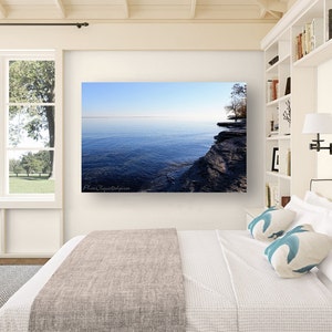 Op de afbeelding: Een foto van een meer met een rotsachtige kustlijn. Het water is diepblauw en de lucht is lichtblauw. De foto hangt aan een witte muur boven een bed met witte beddengoed en blauwe kussens.