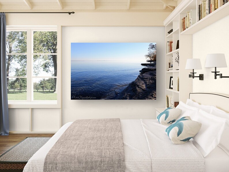 Op de afbeelding: Een foto van een meer met een rotsachtige kustlijn. Het water is diepblauw en de lucht is lichtblauw. De foto hangt aan een witte muur boven een bed met witte beddengoed en blauwe kussens.