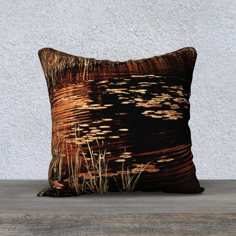 May include: A decorative throw pillow with a black background and a pattern of orange and brown lily pads and reeds reflecting in the water.