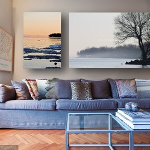 May include: Two framed landscape photographs of a lake with trees and a foggy sky, hanging above a grey sofa with cushions and a glass coffee table with books.
