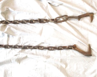 May include: A vintage logging chain with two curved hooks and a series of chain links. The metal is a rusty brown color, suggesting age and use. The chain is laid out on a white fabric background.