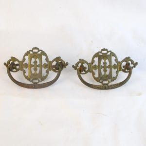 May include: Two antique brass drawer pulls with ornate, decorative designs. Each pull features a central rectangular plate with an intricate pattern, flanked by curved handles. The brass has a weathered, aged appearance, suggesting vintage craftsmanship.