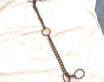 May include: A rusty brown metal chain with three circular rings and a series of chain links. The chain is lying on a white fabric surface. The chain is likely used for restraint or as a decorative element.