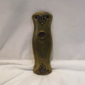May include: A vintage brass door plate with an ornate design. The plate has a keyhole and a round hole for a doorknob.