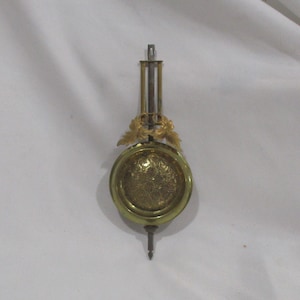 May include: A gold-colored pendulum with a decorative design and a small, pointed tip. The pendulum is suspended from a metal rod.