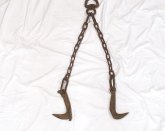 May include: A rusty metal tool with a chain and two curved hooks. The chain is connected to a ring at the top. The hooks are facing opposite directions.