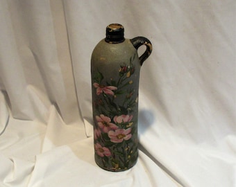 May include: A grey ceramic bottle with a black handle and a floral design painted on the front. The flowers are pink and white with green leaves.