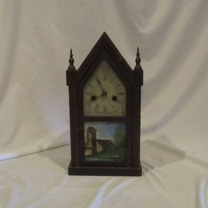 May include: Antique wooden clock with a painted scene of a building and trees. The clock face has Roman numerals and a single hand.