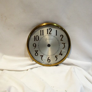 May include: A white clock face with black Roman numeral hour markers and black Arabic numeral minute markers. The clock face has a gold rim and a gold center with the word "Sessions" printed on it. The clock face is made in the U.S.A.