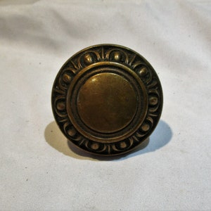 May include: A round brass knob with a decorative design around the edge. The knob has a smooth, circular surface in the center.