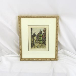 May include: A framed black and white print of a street scene in a European city. The print is in a gold frame with a decorative border.