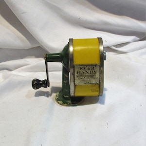 May include: Vintage yellow and green metal pencil sharpener with a crank handle. The sharpener has a label that reads "Ever Handy Pencil Sharpener,  Made in U.S.A."