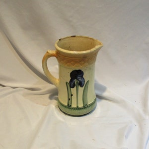 May include: A yellow ceramic pitcher with a blue and green iris flower design. The pitcher has a wide mouth and a handle.