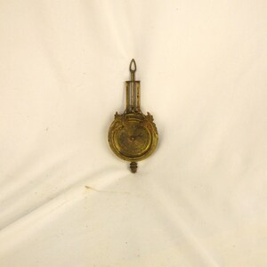 May include: A vintage brass clock pendulum with an ornate design. The pendulum has a circular face with decorative elements and a long, slender suspension rod. The overall color is a warm, aged brass.