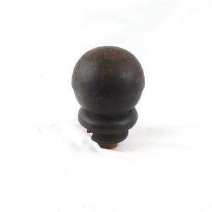 May include: A dark brown wooden finial featuring a spherical top section and a rounded, tiered base. The wood surface displays visible grain, natural wear, and subtle texture variations throughout the rounded form, ending in a small, light-colored attachment point at the bottom.