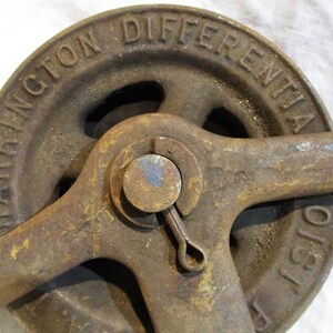 Pu&ograve; includere: Primo piano di un argano differenziale Harrington in ghisa arrugginita. L'argano ha una grande ruota con il testo "Harrington Differential Hoist" in rilievo sul lato. L'argano ha un braccio di leva e una piccola maniglia.