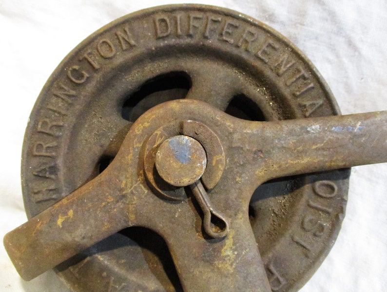 Pu&ograve; includere: Primo piano di un argano differenziale Harrington in ghisa arrugginita. L'argano ha una grande ruota con il testo "Harrington Differential Hoist" in rilievo sul lato. L'argano ha un braccio di leva e una piccola maniglia.