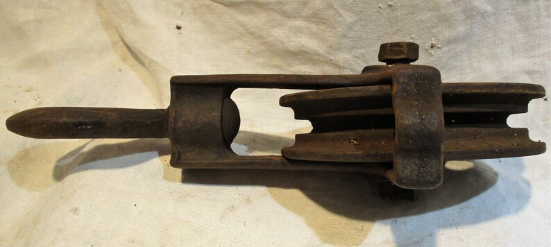 May include: A rusty metal pulley with a single wheel and a handle. The pulley has a metal loop on the side for attaching a rope.