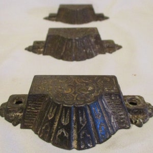 May include: Three antique cast iron drawer pulls with ornate designs. The pulls are dark brown with gold accents and have a scalloped edge.