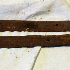 May include: Two rusty metal hinges, each with a rounded end and several holes. The hinges are lying on a white fabric surface.