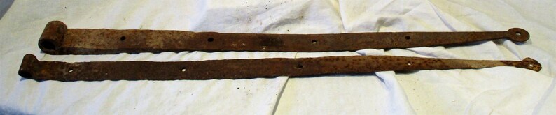 May include: Two rusty metal hinges, each with a rounded end and several holes. The hinges are lying on a white fabric surface.