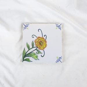 May include: Square white tile with a hand-painted floral design. The yellow and orange flower is accented with green leaves and black stems. Blue and green corner accents complete the design.