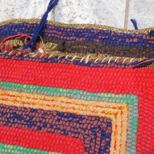 May include: Close-up of a colorful woven rug with a red, blue, green, and brown pattern. The rug has a frayed edge and is made of thick yarn.