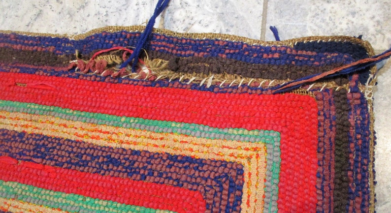 May include: Close-up of a colorful woven rug with a red, blue, green, and brown pattern. The rug has a frayed edge and is made of thick yarn.
