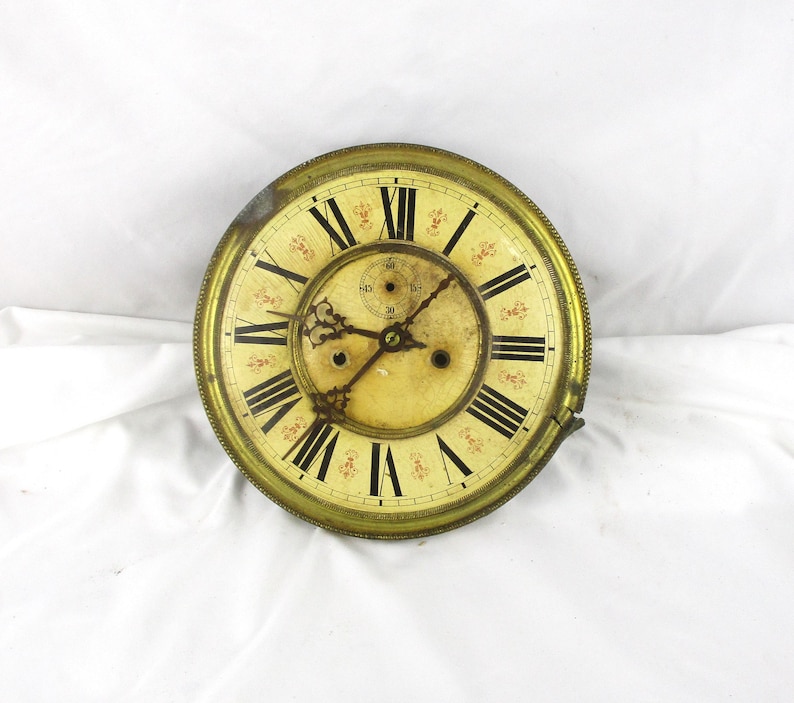 Peut inclure: Cadran d'horloge ancien avec une lunette en laiton et un cadran cr&egrave;me. Les chiffres romains noirs indiquent les heures, avec des motifs floraux d&eacute;licats. Le cadran montre des signes d'&acirc;ge et d'usure, ajoutant &agrave; son charme vintage.