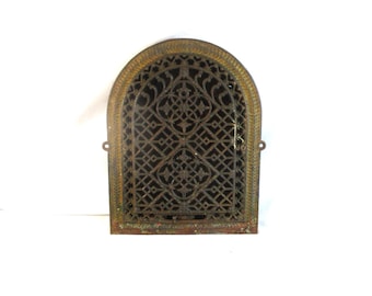 May include: An antique, arched metal vent cover with an ornate, geometric lattice design. The frame has a bronze finish with a decorative border. Two small loops are on the sides for mounting. The vent cover is likely for a fireplace or heating system.