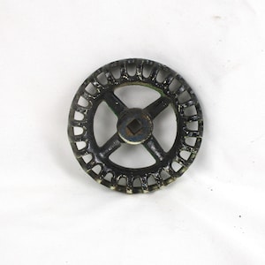 May include: A circular, black metal wheel with a series of small, open rectangles around the outer edge. The wheel has four spokes that meet in the center, with a square opening. The wheel has a distressed, vintage appearance.
