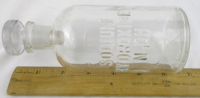 Może przedstawiać: Przezroczysta szklana butelka z korkiem, oznaczona napisem "SODIUM HYDROXIDE N.OH". Butelka leży poziomo na drewnianej linijce. Linijka jest oznaczona w centymetrach.