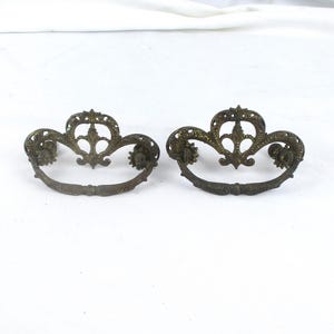 May include: Two matching antique brass drawer pulls featuring an ornate, openwork backplate with a central fleur-de-lis motif and a curved, swinging bail handle. The metal has a dark, aged patina with visible texture and intricate detailing.