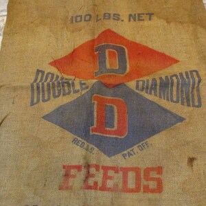 May include: A vintage burlap sack with the text "100 LBS. NET" printed at the top. The sack features a large, red and blue diamond logo with the letters "D D" in the center. The text "DOUBLE DIAMOND FEEDS" is printed below the logo.