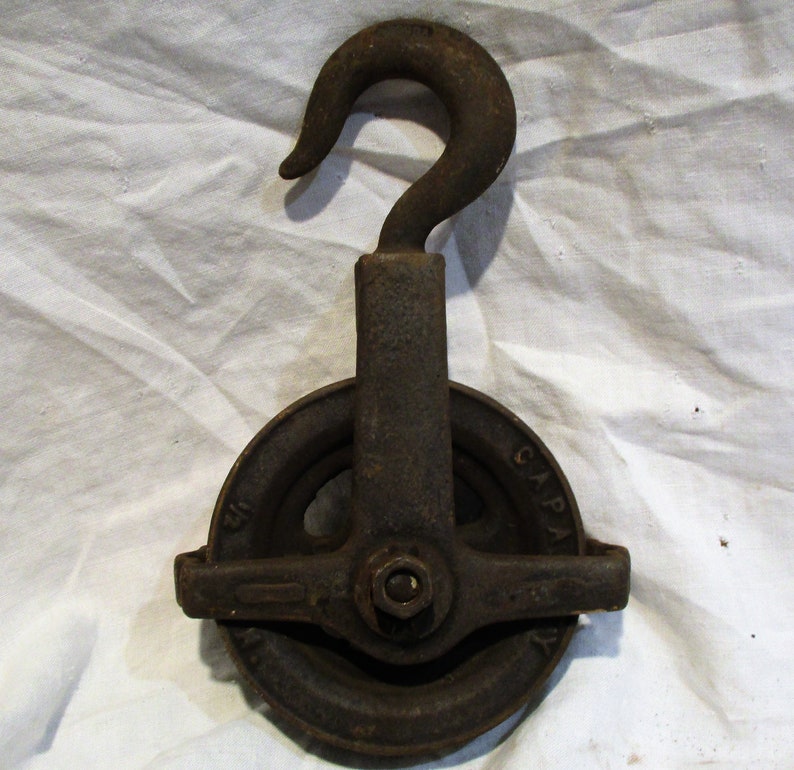 May include: A vintage cast iron pulley with a hook and a wheel with a groove for rope. The pulley is marked "CAPA" on the side.