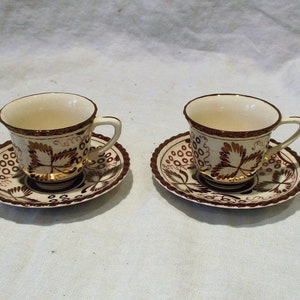 May include: Two white teacups and saucers with a brown and gold floral design. The cups and saucers have a scalloped edge.